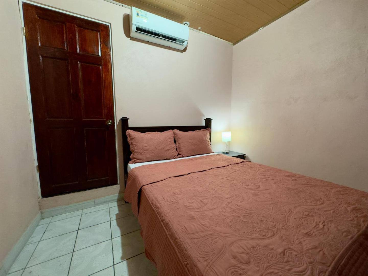 A simple room with a double bed with a rust-colored bedspread, a wooden door and an air conditioner on the wall